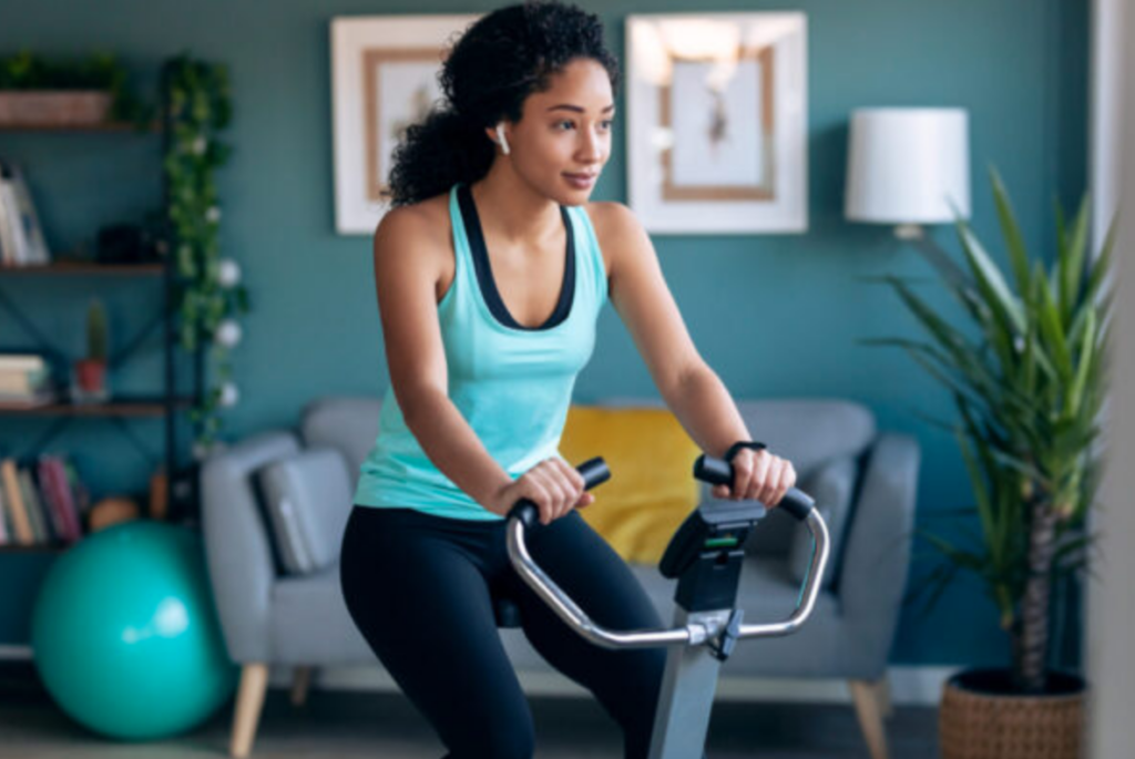 Shot of sporty african young woman exercising on smart stationary bike and listening to music at home.